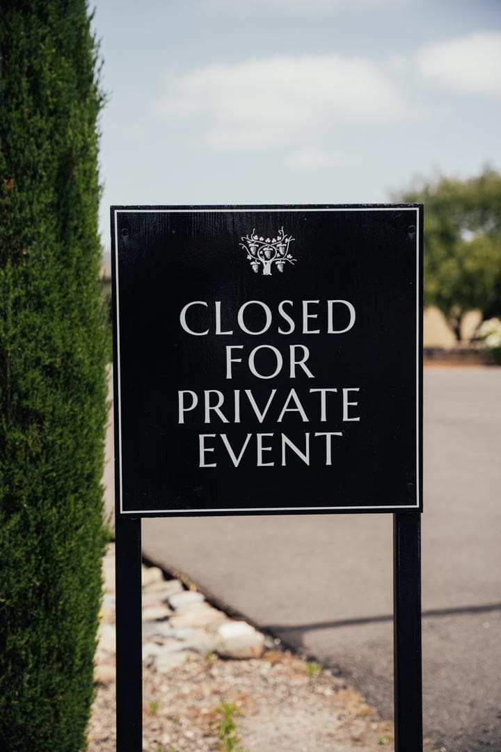 A black sign with white text reads "Closed for Private Event" standing on a paved area with trees and shrubs in the background.