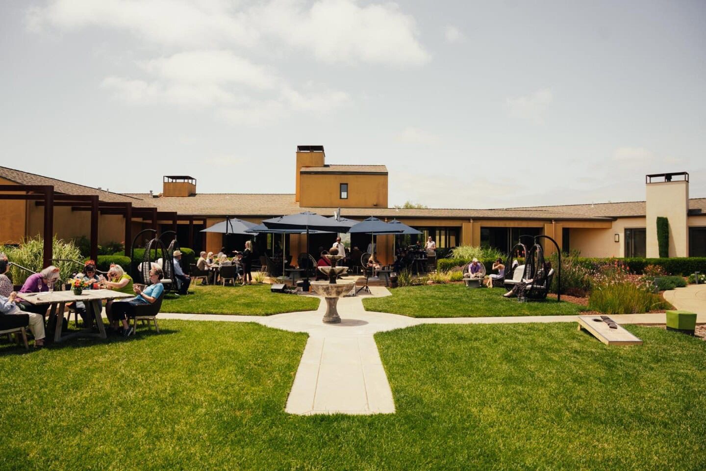 People are seated at tables and under umbrellas in a landscaped courtyard outside a large building on a sunny day.