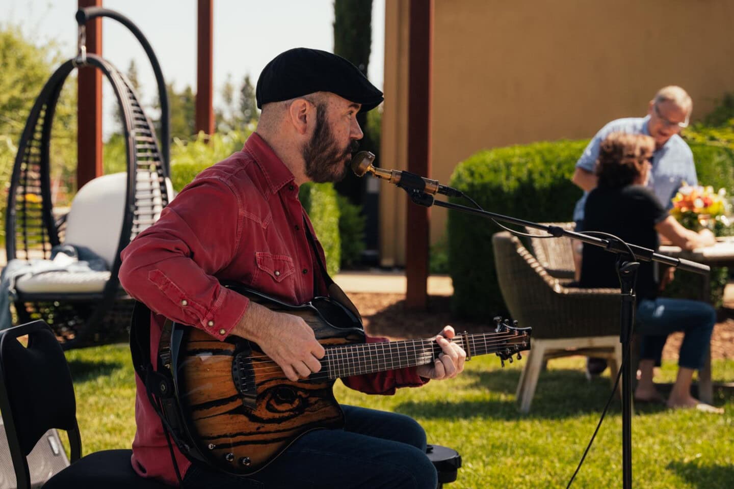 A man in a red shirt and black cap plays an acoustic guitar and sings into a microphone outdoors; two people are seated at a table in the background.