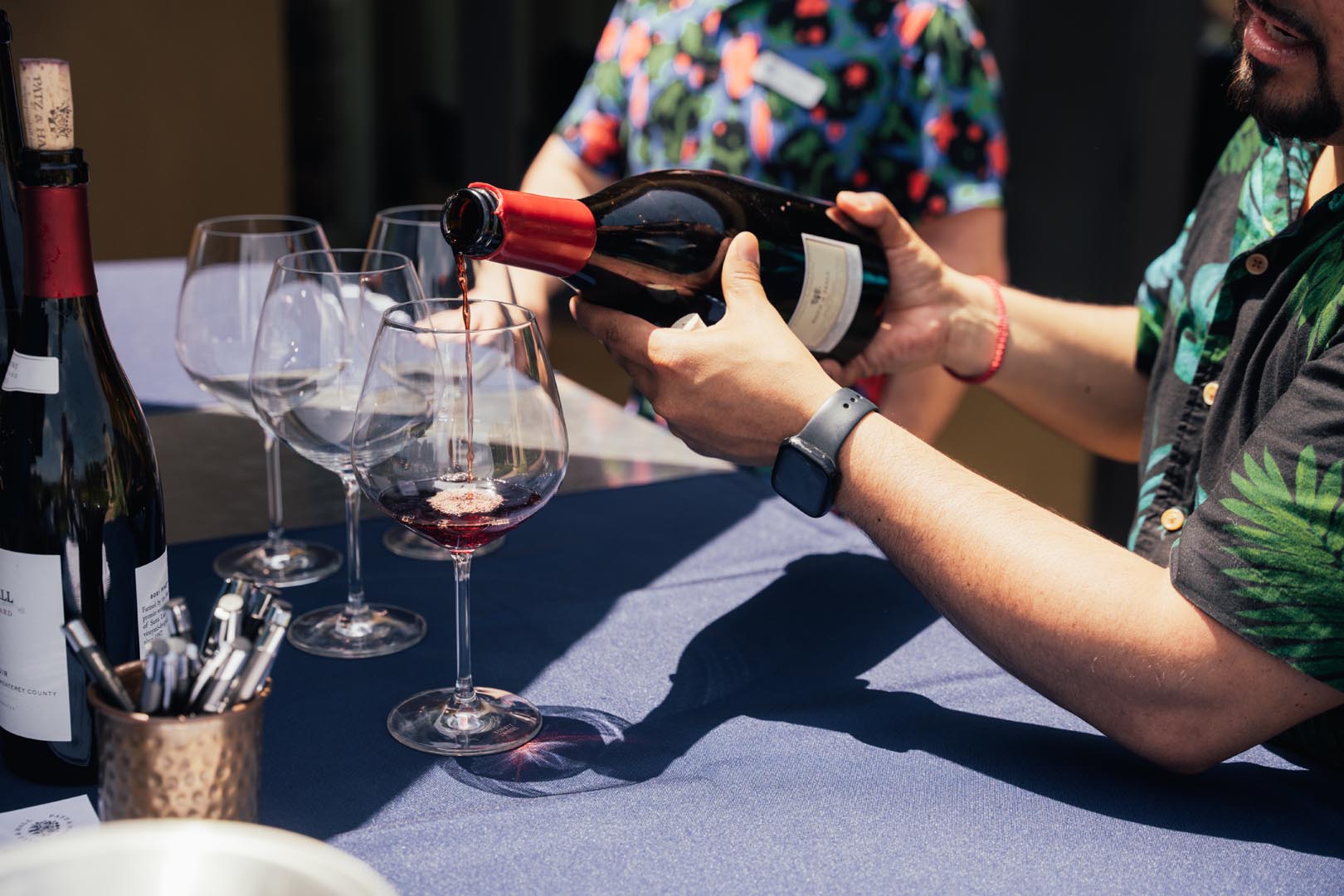 A person pours red wine into glasses on a table covered with a blue tablecloth, with another person standing nearby and several bottles and pens visible.