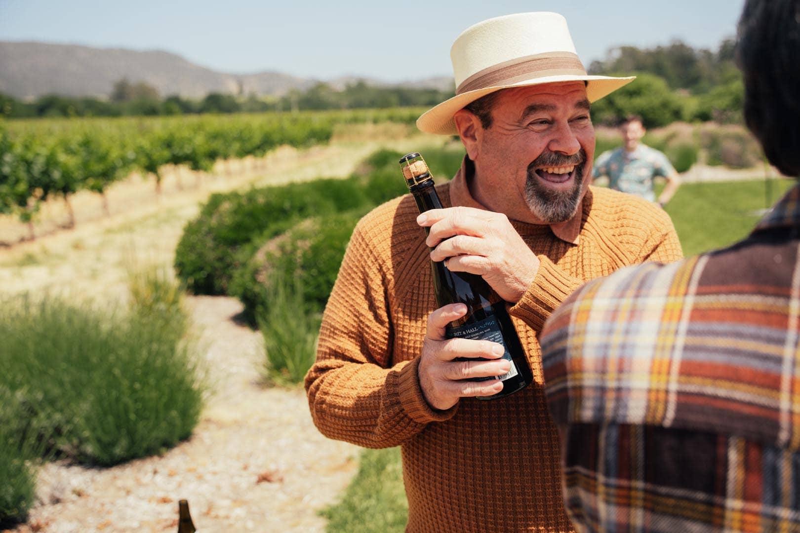A man wearing a hat and brown sweater smiles while holding a wine bottle outdoors in a vineyard on a sunny day.