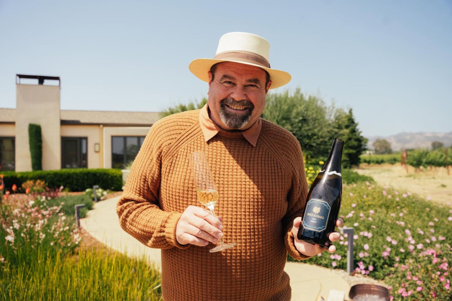A man in a brown sweater and hat stands outside holding a wine bottle and a glass of white wine, with a garden and house in the background.