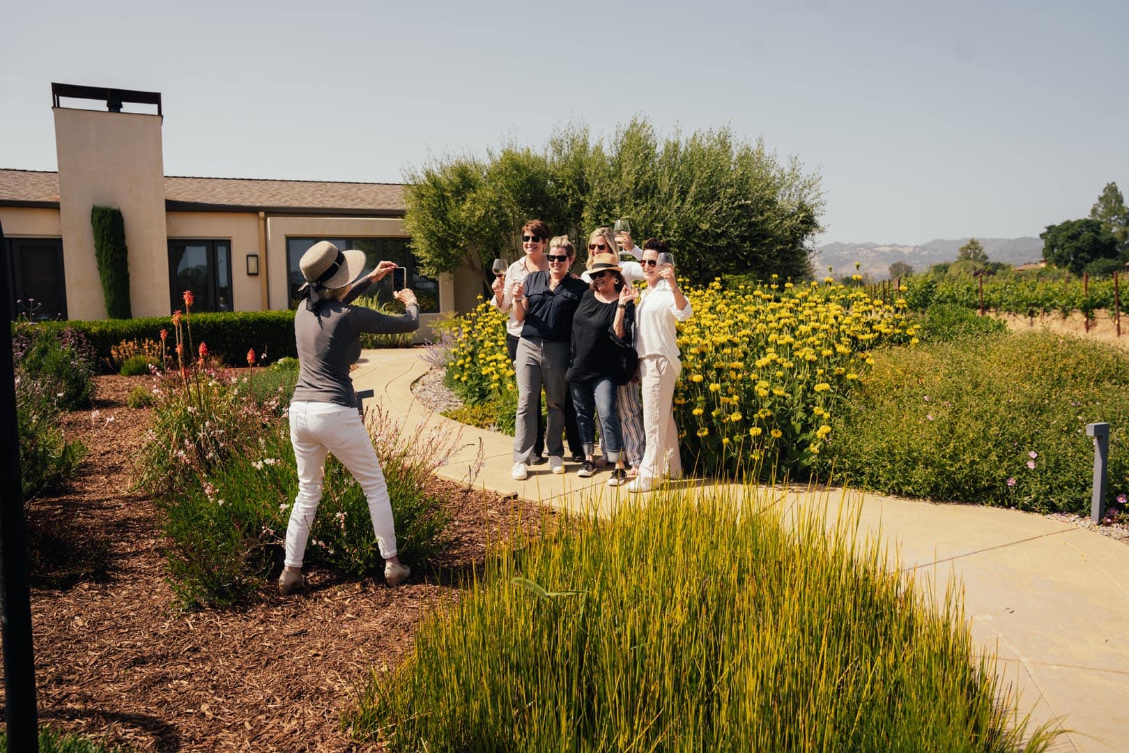 A person takes a photo of a group of six people posing together on a garden path surrounded by greenery and yellow flowers near a building.