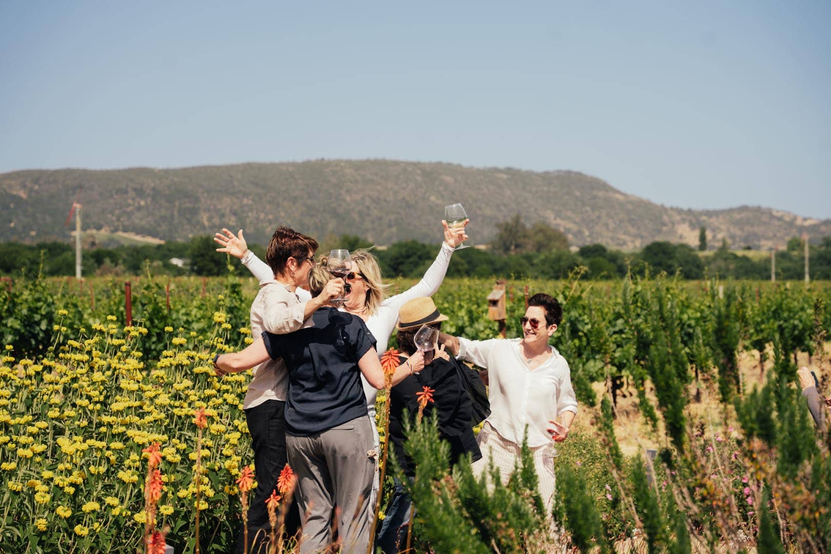 A group of people standing in a vineyard with flowers, smiling and laughing together, one person holding up a wine glass, with hills in the background.