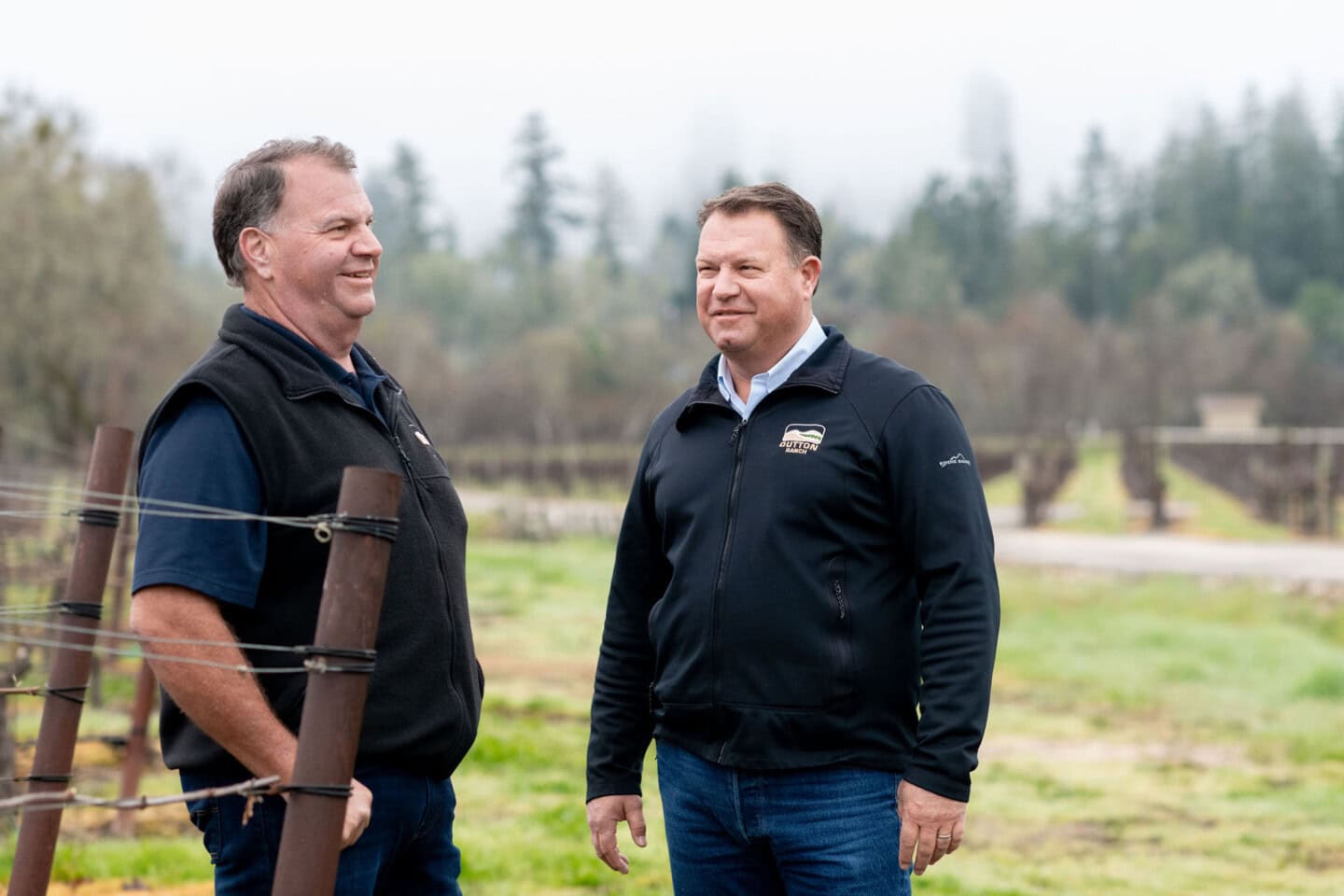 Two men stand and converse beside a vineyard on a cloudy day, wearing casual jackets and jeans, with rows of grapevines and trees in the background.