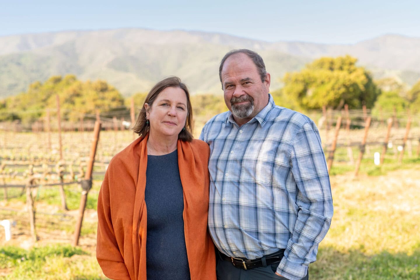 A woman and a man stand side by side in a vineyard with mountains and trees in the background on a clear day.