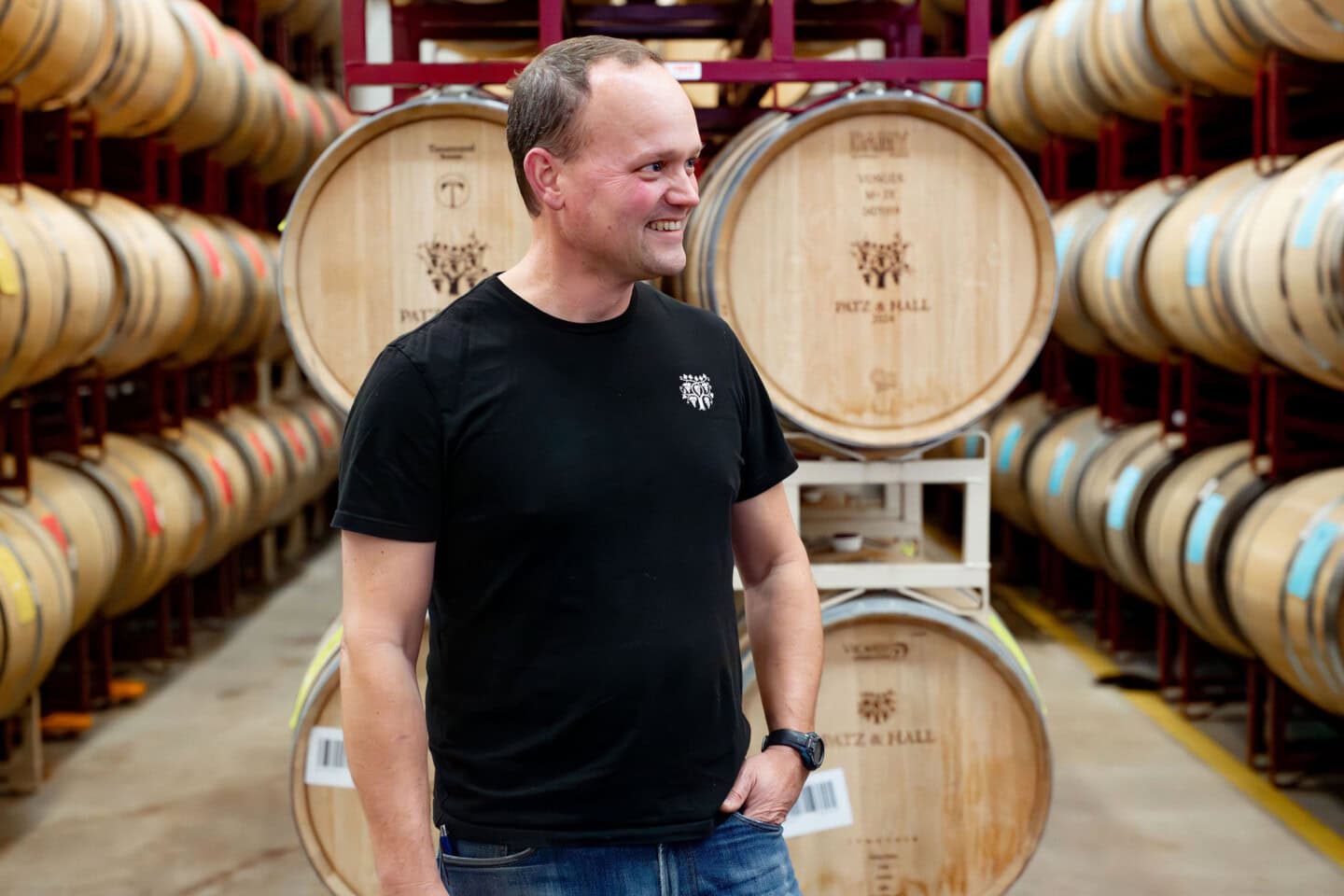A man in a black t-shirt stands smiling in a wine cellar, surrounded by stacked wooden barrels.
