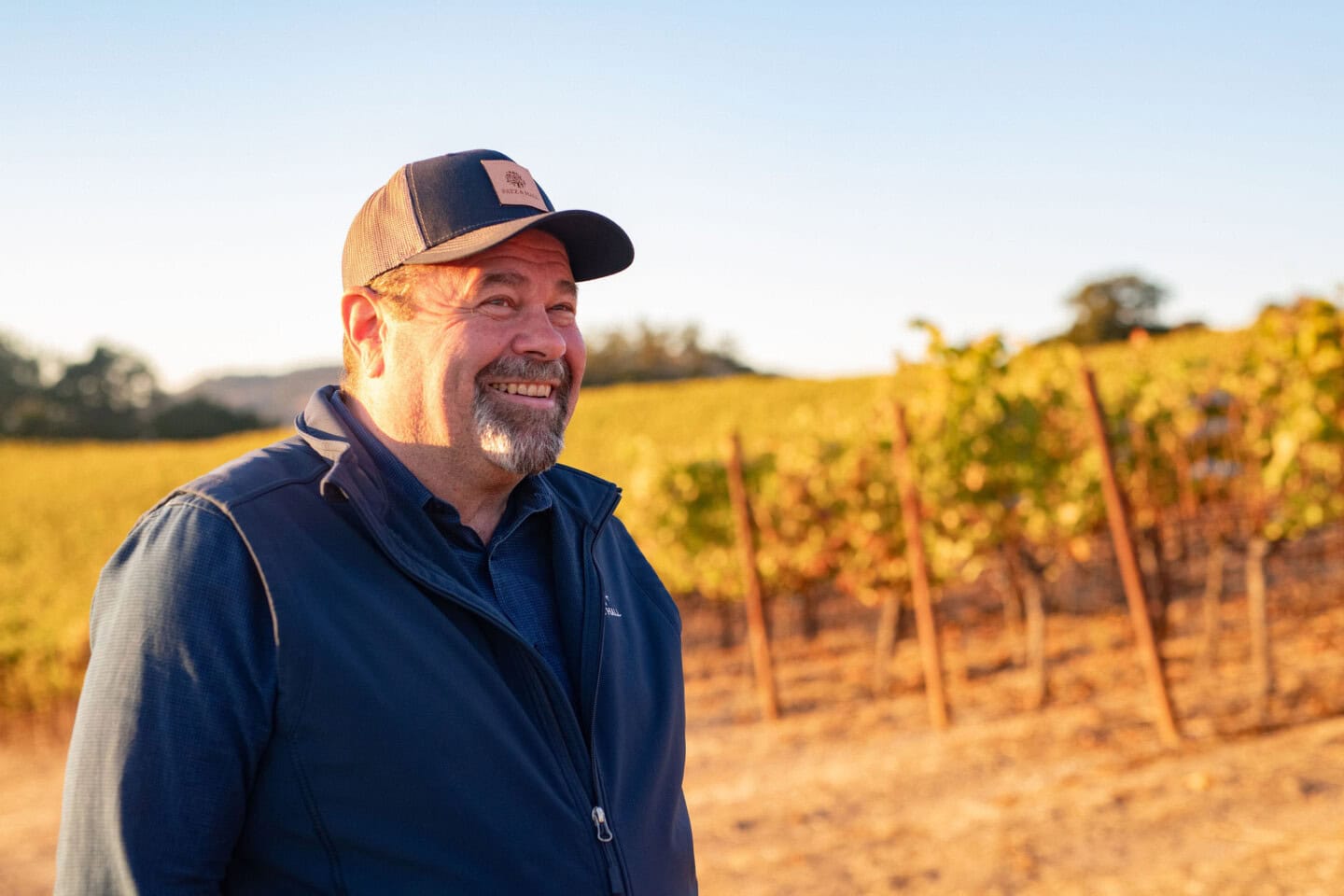 A man in a blue jacket and baseball cap stands smiling in a vineyard during daylight. Rows of grapevines and clear sky are visible in the background.