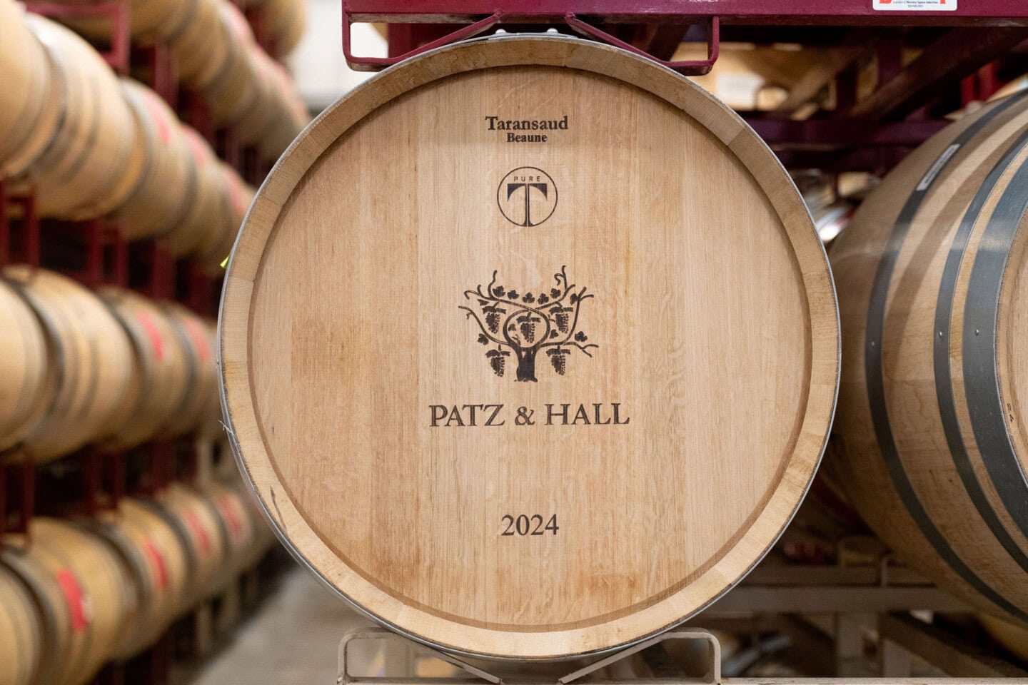 A wooden wine barrel labeled "Patz & Hall 2024" is stored on a rack among other barrels in a cellar.