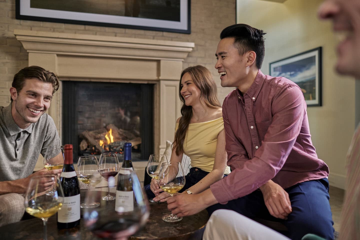 Four people sit around a table with wine glasses and bottles, smiling and talking in a cozy room with a lit fireplace in the background.
