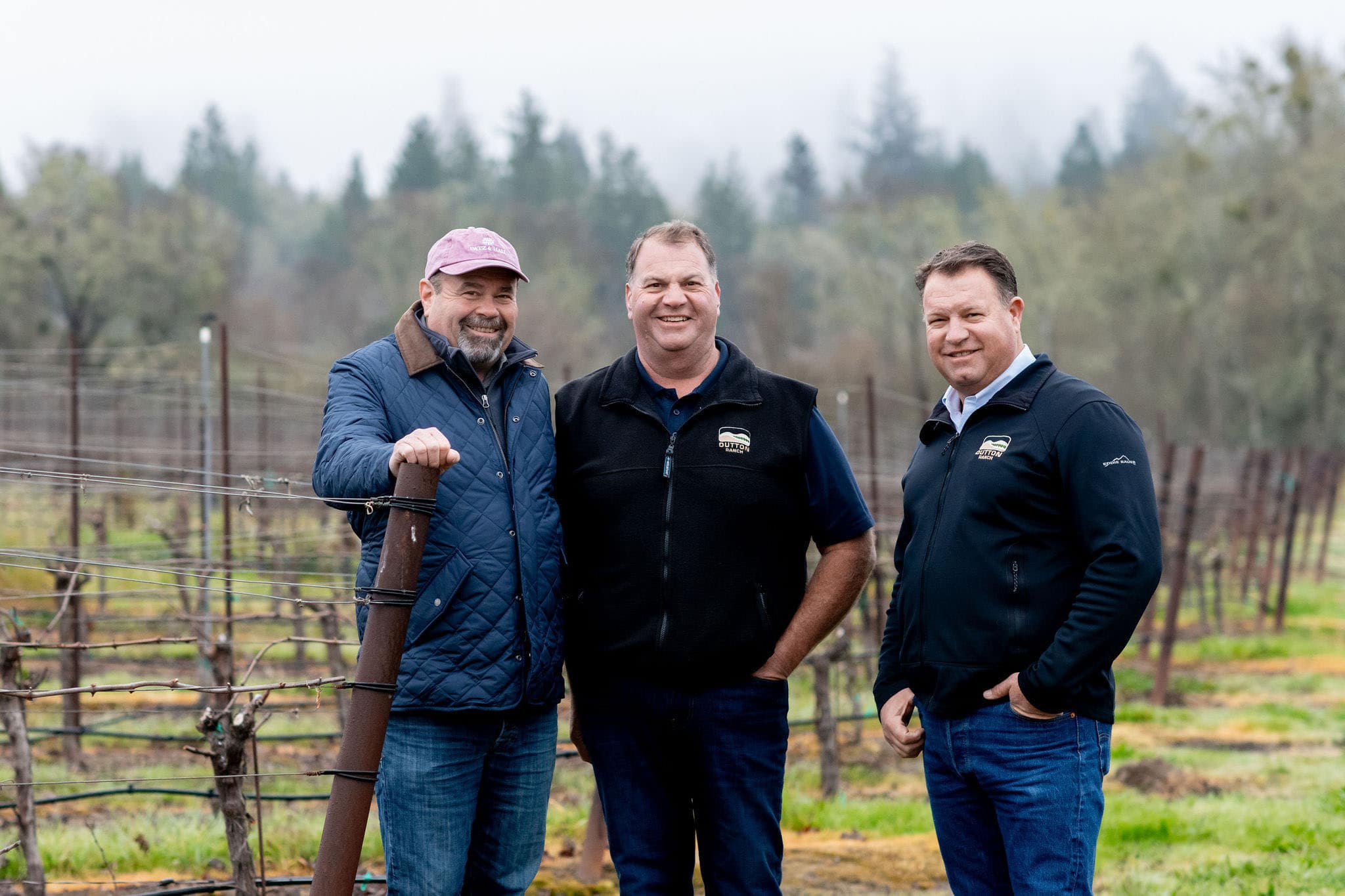 Three men stand together in a vineyard on a cloudy day, with leafless vines and trees visible in the background.