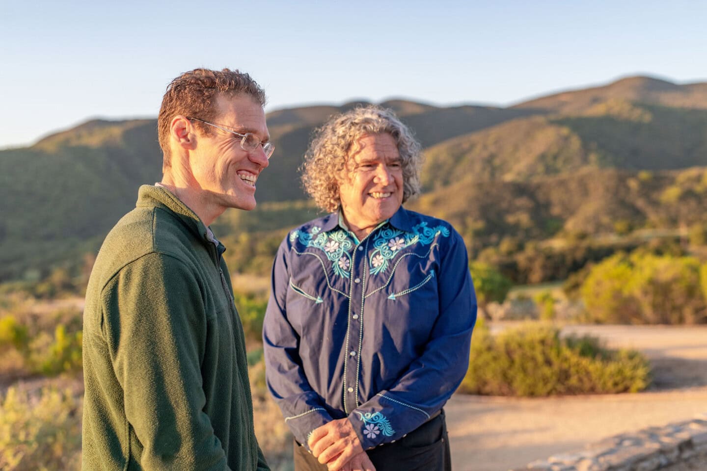 Two men standing outdoors, smiling, with mountains and greenery in the background under a clear sky.