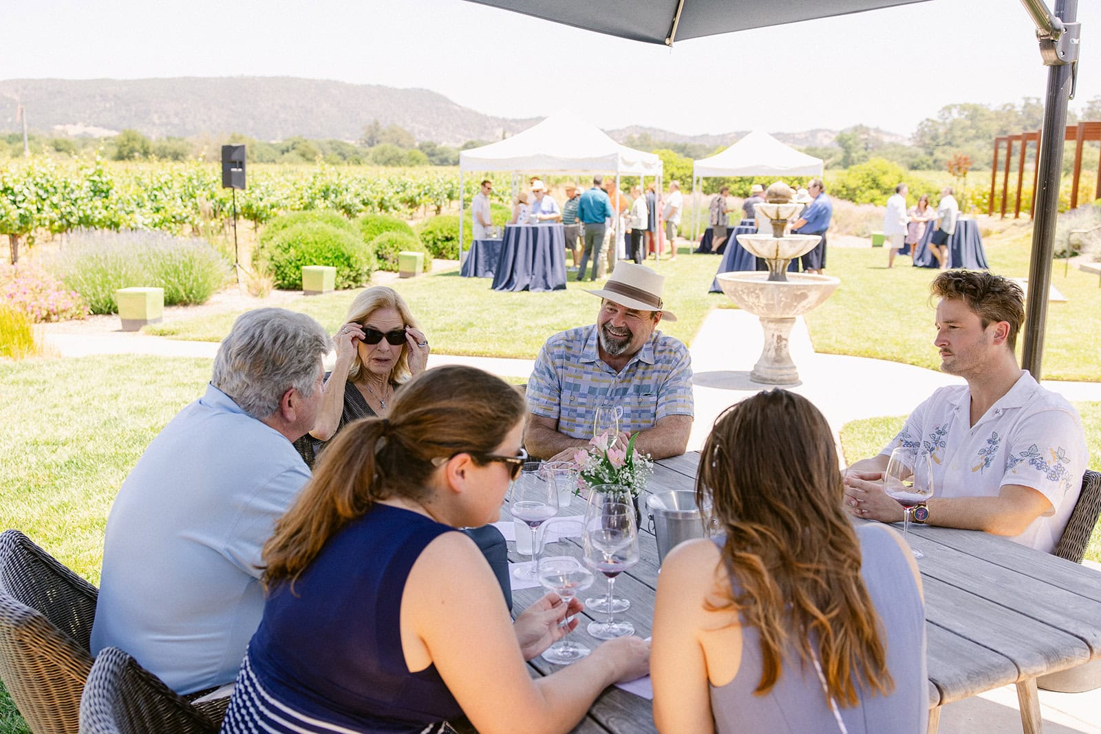 A group of people sit around a wooden table outdoors at a vineyard, talking and drinking wine, with other guests and white tents visible in the background.