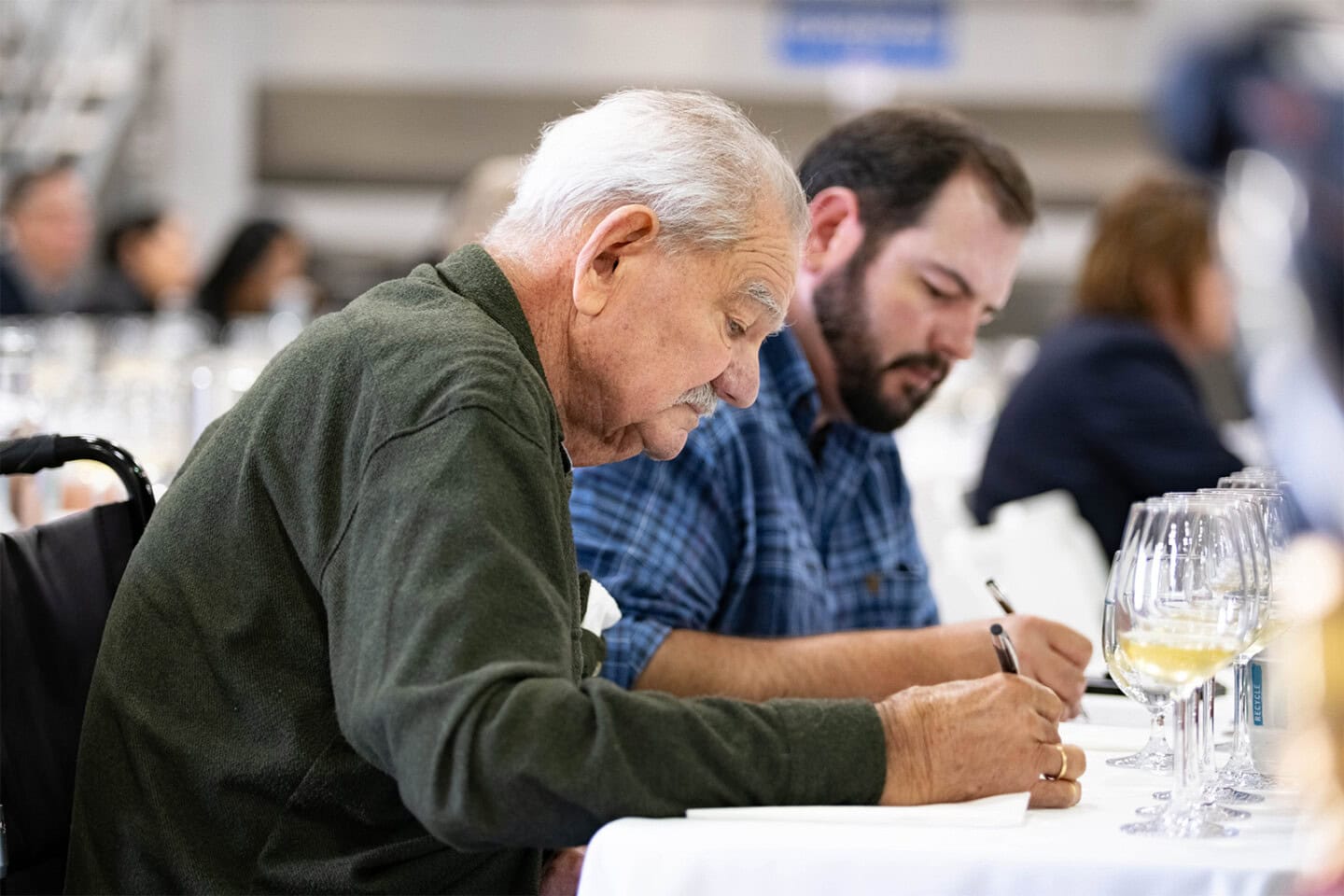 Two men sitting at a table write on paper, with wine glasses in front of them, in a well-lit conference or seminar setting.